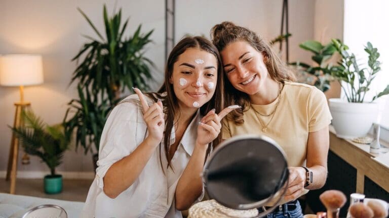 2 chicaas cuidándose la piel con cremas, sonriendo frente al espejos