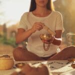 Mujer practicando meditación con un cuenco tibetano, rodeada de elementos de relajación en un ambiente cálido y natural.