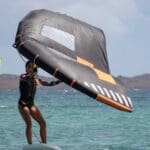 Persona practicando WingFoil en aguas cristalinas de Playa Blanca, Lanzarote. Tabla con hidroala y vela, deslizándose sobre el mar con un paisaje espectacular de fondo. Deportes acuáticos emocionantes en un entorno paradisíaco.