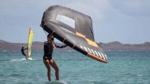 Persona practicando WingFoil en aguas cristalinas de Playa Blanca, Lanzarote. Tabla con hidroala y vela, deslizándose sobre el mar con un paisaje espectacular de fondo. Deportes acuáticos emocionantes en un entorno paradisíaco.