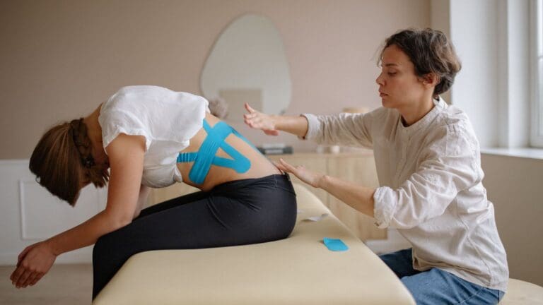 Fisioterapeuta aplicando vendaje neuromuscular en la espalda de una paciente para aliviar el dolor y mejorar la movilidad en una sesión de terapia.