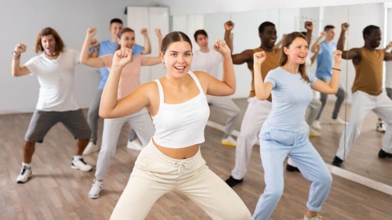 Grupo de personas disfrutando una clase de baile en una academia, realizando movimientos coordinados con entusiasmo y energía en un salón con espejos.