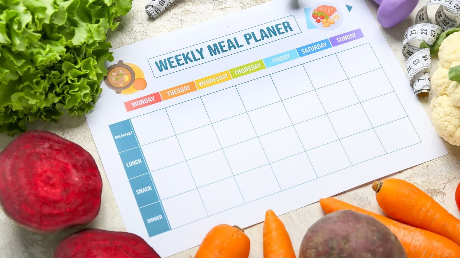Mesa con alimentos saludables, frutas frescas y un planificador semanal de comidas, representando un estilo de vida con alimentación equilibrada.