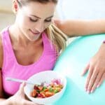 Mujer deportista sonriente con ropa deportiva sostiene un bol de ensalada fresca tras el entrenamiento junto a un balón de ejercicio, ilustrando hábitos de alimentación saludable para mejorar el rendimiento y evitar errores comunes en la nutrición deportiva.