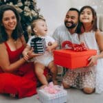 Familia sonriendo junto al árbol de Navidad mientras intercambian regalos para niños