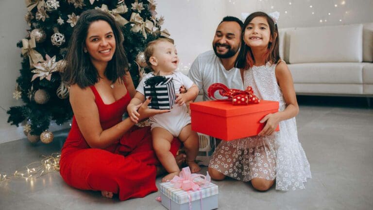 Familia sonriendo junto al árbol de Navidad mientras intercambian regalos para niños