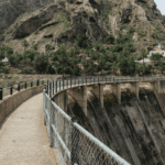 Vista de presa y pasarela en zona rural montañosa con casas rodeadas de vegetación en España.