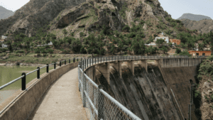 Vista de presa y pasarela en zona rural montañosa con casas rodeadas de vegetación en España.
