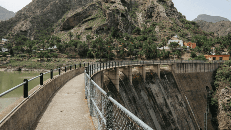 Vista de presa y pasarela en zona rural montañosa con casas rodeadas de vegetación en España.