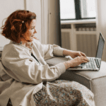 Mujer sonriente utilizando un portátil desde el sofá mientras realiza formación online sobre hábitos saludables desde casa.