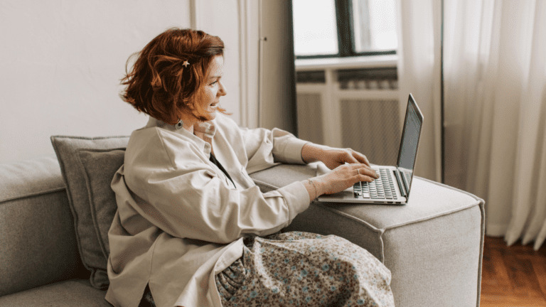 Mujer sonriente utilizando un portátil desde el sofá mientras realiza formación online sobre hábitos saludables desde casa.